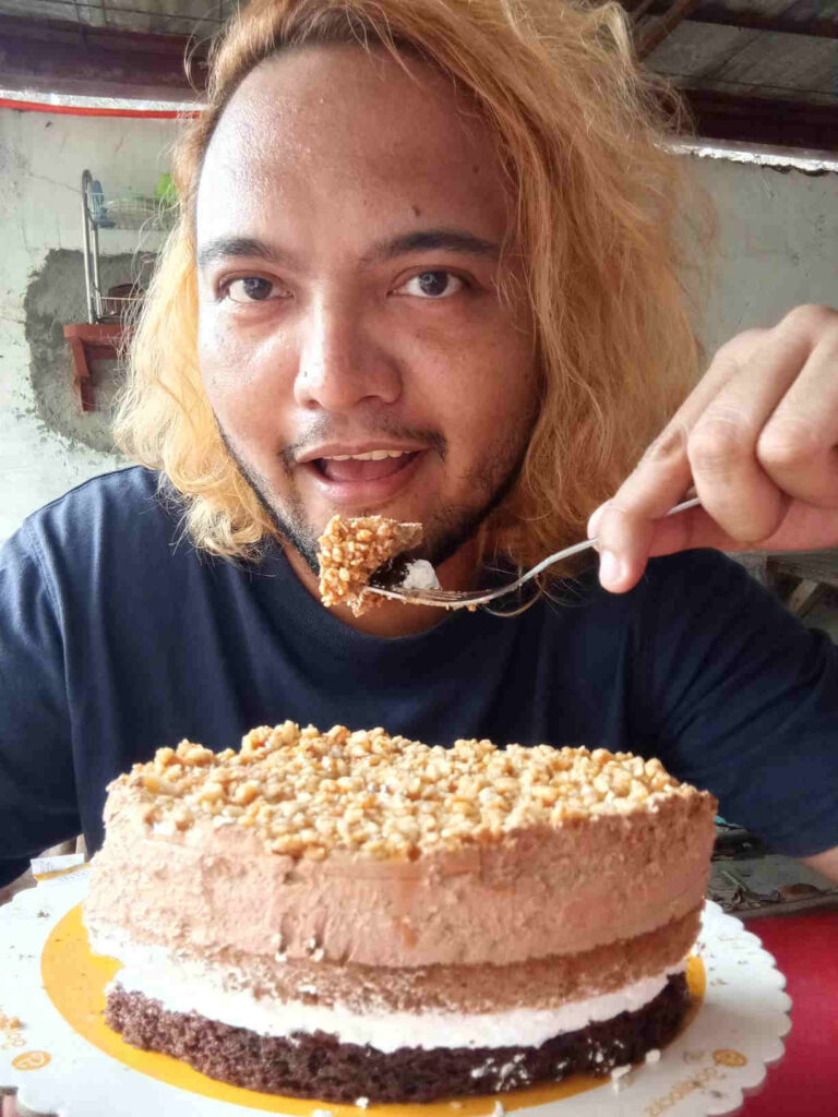 Blonde Filipino male eating Coffee Layered Cake by Goldilocks Bakeshop using a fork with his left hand.
