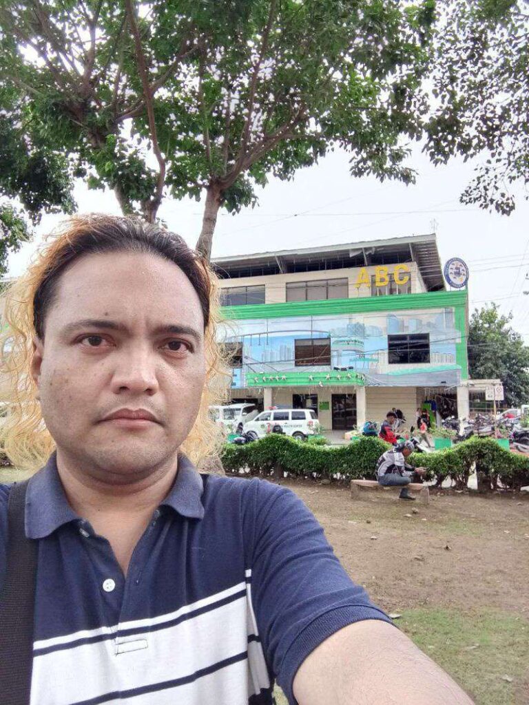 A selfie of a person with shoulder-length light brown hair, wearing a navy blue and white striped polo shirt. The person is standing outdoors in front of a building with the letters "ABC" visible. The building has a mural on its side and is surrounded by greenery and a few people are visible sitting on a bench in the background. The overall setting appears to be a park or public area.