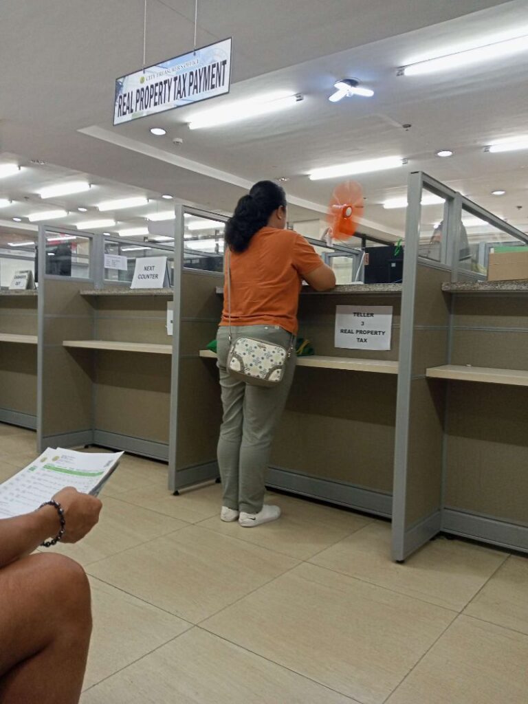 A photo of the interior of a Real Property Tax Payment office. A woman with long dark hair, wearing an orange shirt and light-colored pants, stands at a counter. The counter is divided into sections by cubicle-like partitions. A small desk fan sits on the counter. Another person's legs and hand holding paperwork are visible in the bottom left corner. The overall setting is clean and brightly lit.