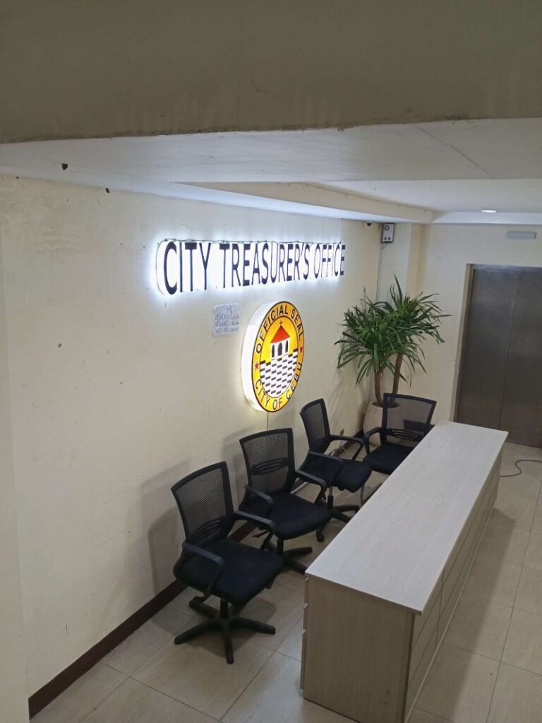 An overhead shot of a City Treasurer's Office waiting area. The image shows a light beige reception desk, four black office chairs, a lit sign reading "CITY TREASURER'S OFFICE," and a circular illuminated emblem on the wall. A potted plant is also visible. The overall setting is clean and orderly.