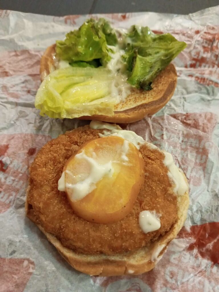 A slice of lettuce on the top bun and a slice of tomato on top of fried chicken fillet on the bottom bun.