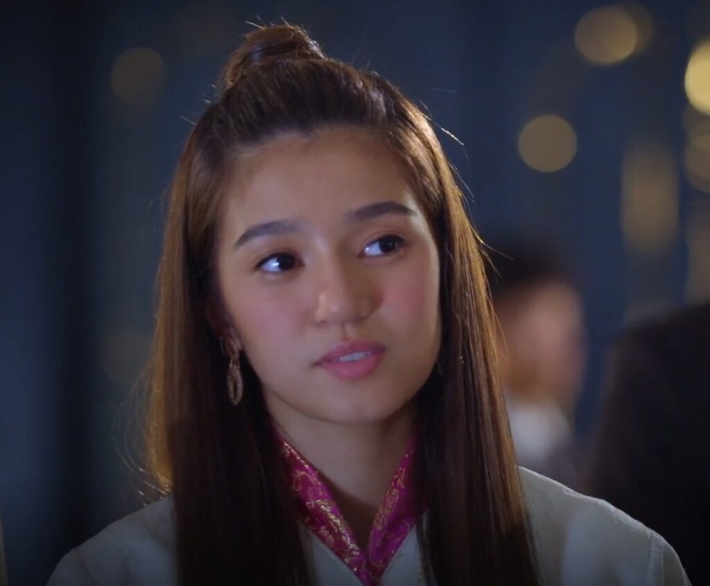 Close-up shot of a young woman with long brown hair styled in a half-updo. She's wearing a light-colored, possibly off-white or beige, garment with a fuchsia or magenta patterned collar. The background is blurred but shows a dimly lit indoor setting with other people out of focus. The woman has a thoughtful or pensive expression.