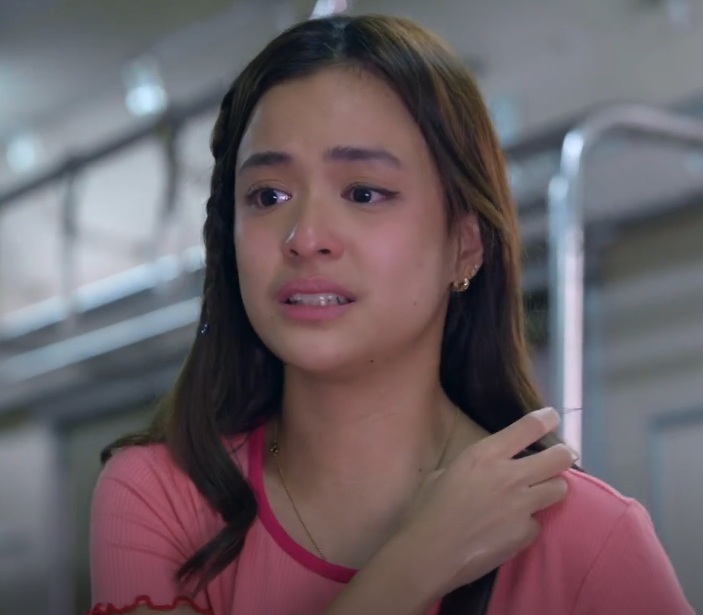 Close-up shot of a young woman with long brown hair, wearing a light pink shirt. She appears to be upset or distressed, with tears in her eyes and a worried expression. Her hand is lightly touching her shoulder. The background is blurred, suggesting an indoor location.