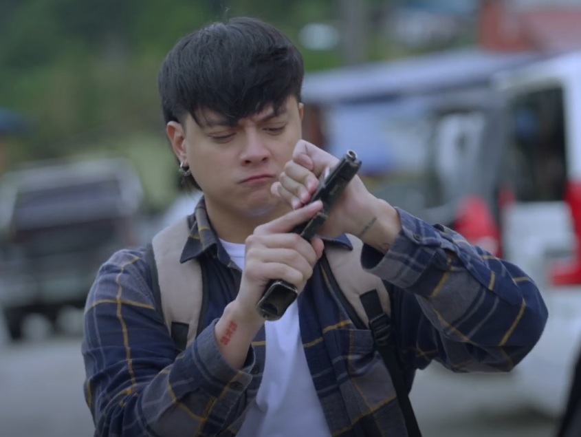 A young man with short dark hair is shown outdoors, carefully examining or manipulating a handgun. He's wearing a plaid shirt and a backpack. The background is blurred, showing parked cars and a suggestion of a street scene. The overall impression is one of tension or suspense.