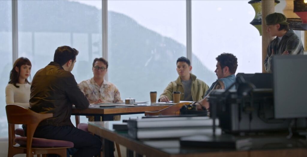 A group of five young adults are seated around a table in a modern office or meeting room with large windows offering a view of mountains in the distance. One person stands near a window, arms crossed. The group appears to be in a meeting or discussion; some have coffee cups on the table. The atmosphere seems professional but relaxed. Part of another table or desk with equipment is in the foreground, slightly out of focus.