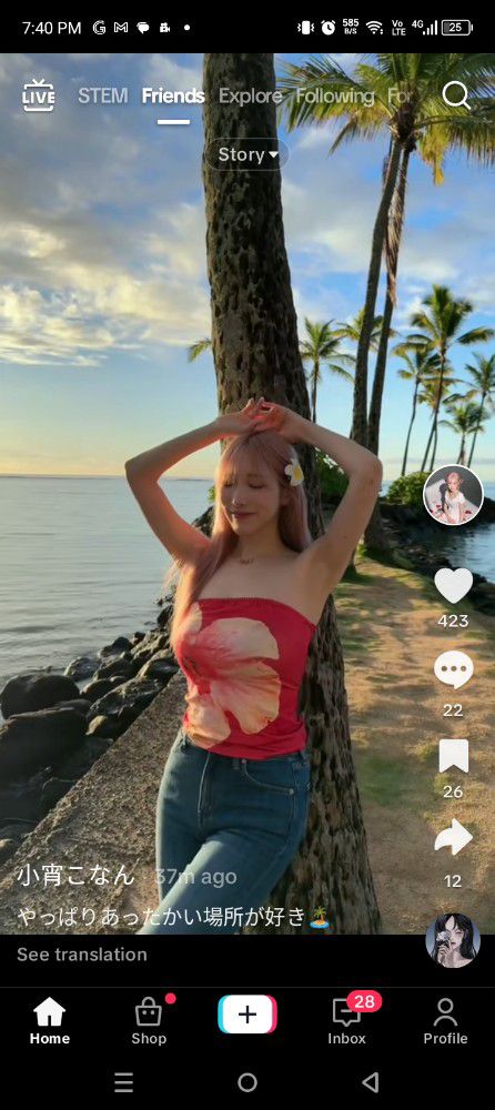 A phone screen shows a social media post. A young woman with long pink hair, wearing a red hibiscus-print tube top and jeans, poses against a tree trunk on a beach at sunset. Palm trees line the beach. The post includes a like counter (423), comment counter (22), a bookmark (26), a share counter (12), and a caption in Japanese that translates to something like, "I like places with a nice atmosphere." The phone's bottom navigation bar shows icons for Home, Shop, Create, Inbox, and Profile.