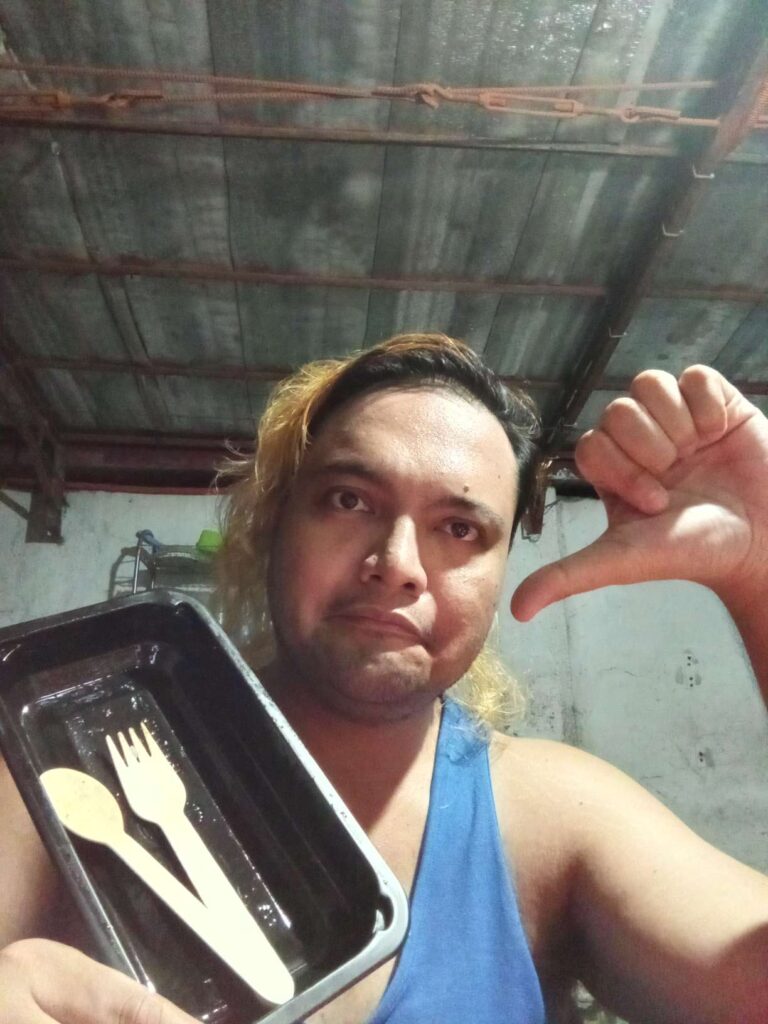 Robin Laviña gives a thumbs down to Uncle Brew pork sisig meal. He is eating in the outdoor kitchen and showing the empty black food container with wooden spoon and fork placed inside.