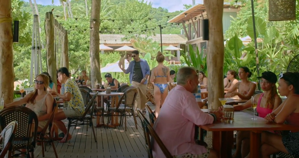 A wide shot of an outdoor restaurant or bar, possibly on a beach or tropical location. Numerous people are seated at various tables, enjoying drinks and conversation. Some are dressed in swimwear, while others are in casual attire. A man in a dark shirt and khaki pants is walking through the scene. The setting is lush and green, with wooden structures and tropical plants visible. The overall atmosphere is relaxed and social.