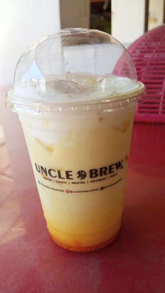 Mango Latte from Uncle Brew in a clear plastic cup and cover. The background shows the drink is on top a red table and behind is a blurred view of white concrete and tiles.