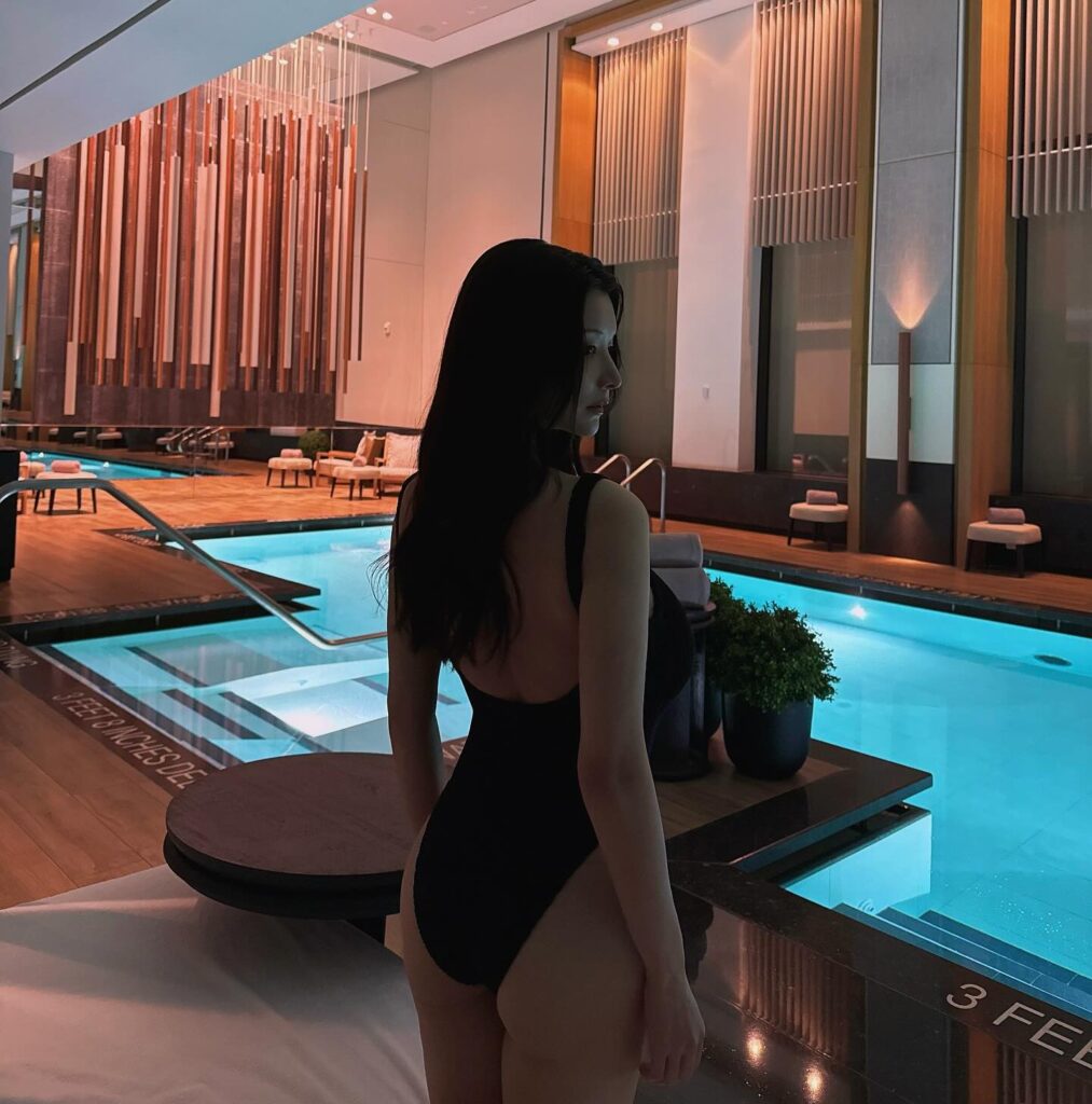 Japanese pornstar Nagi Hikaru hows her sexy back in a black one piece swimsuit that accentuates her back, She's showing the side view of face and chest. Her long black hair flows in her back and the length is up to the chest region. The place looks like an indoor swimming pool of a hotel. The hotel pool looks very modern with a wooden interior design accent.